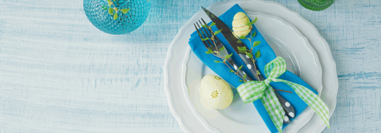 Tavola di Pasqua apparecchiata con piatti bianchi, tovagliolo azzurro, posate decorate con rametti e uova colorate